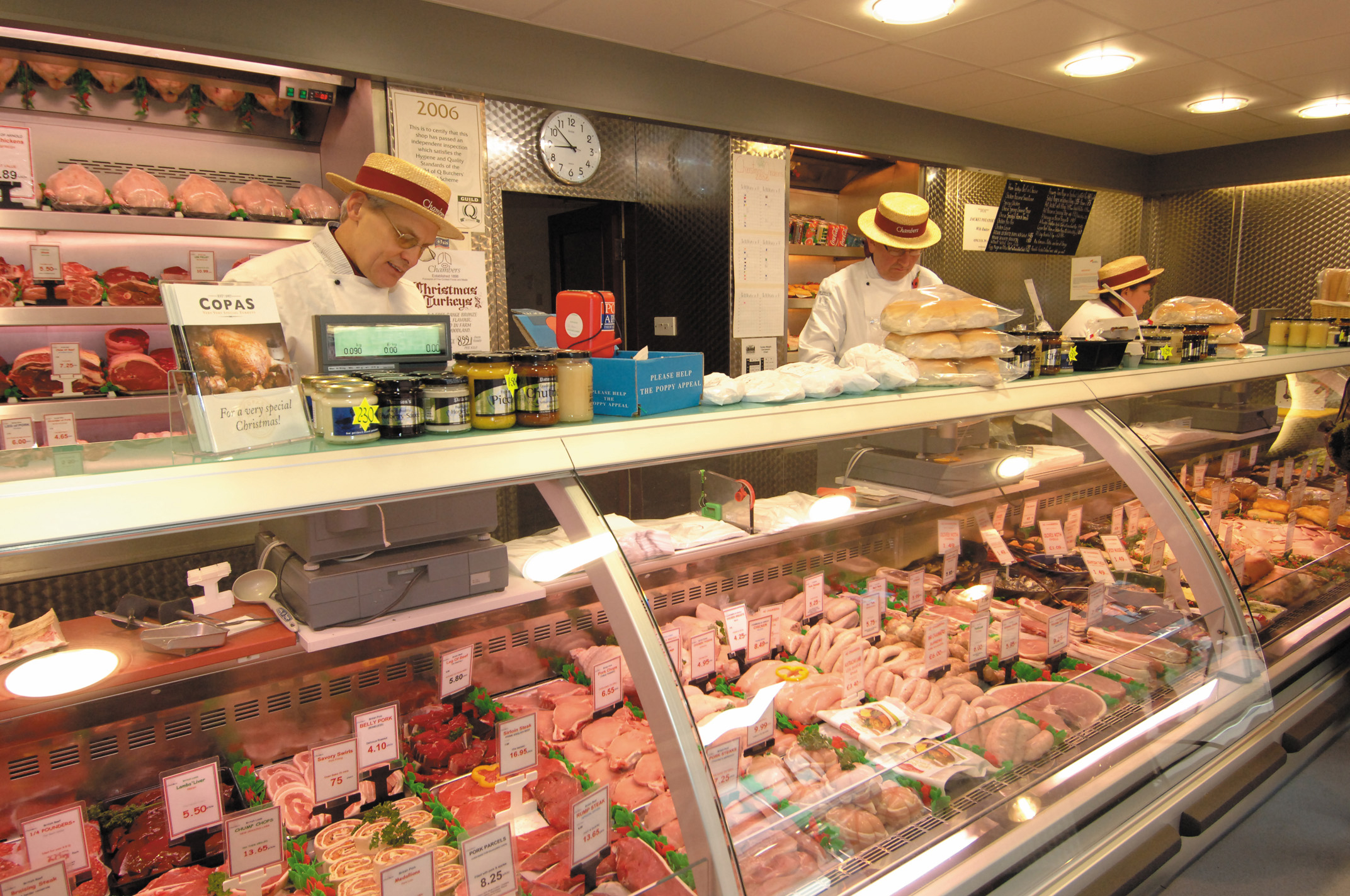 Two Chambers butchers in white aprons and traditional straw boater hats working behind the glass counter at 31 Front Street